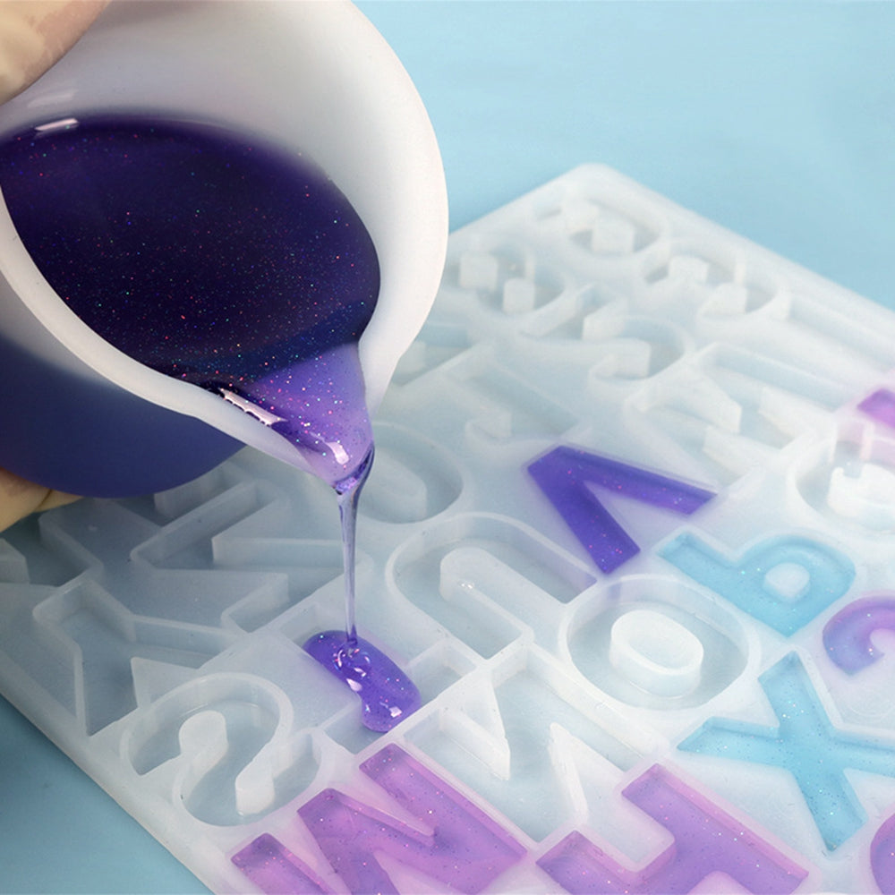Silicone graduated measuring cup pouring purple liquid into a silicone letter mold.